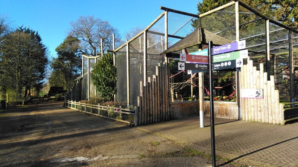 Entrance to a zoo with directional signs pointing towards various animal exhibits, surrounded by trees and winter scenery.